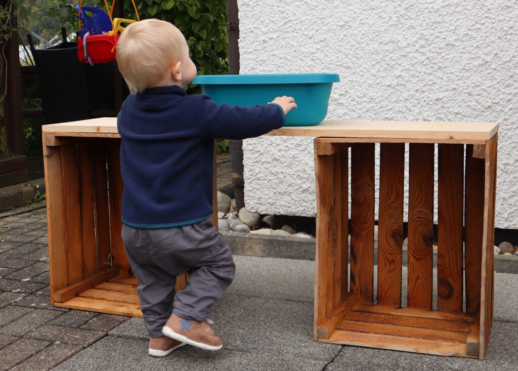Das Foto zeigt ein Kind, das neben zwei Holzkisten steht auf denen ein Brett liegt