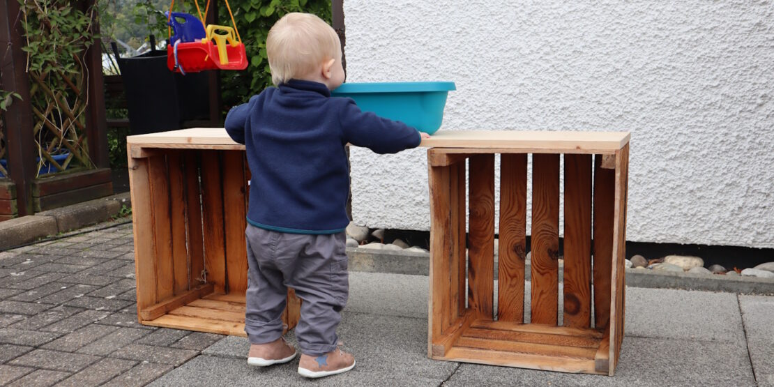 Das Foto zeigt ein Kleinkind neben zwei Obstkisten, auf denen ein Brett liegt