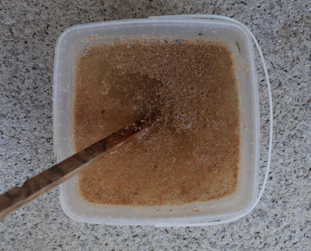 Das Foto zeigt einen Eimer mit braunem Wasser in dem Krümel schwimmen. Ein Holzstab steckt im Eimer.