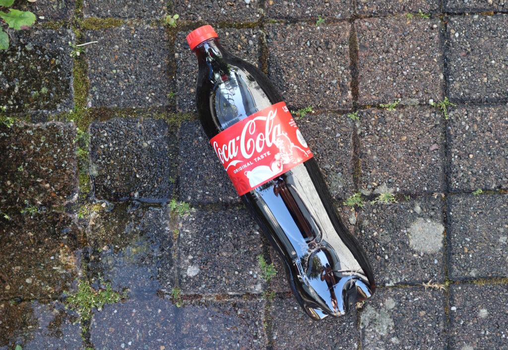 Das Foto zeigt eine Flasche Cola auf einem Steinboden. In den Steinzwischenräumen erkennt man Grünbelag.