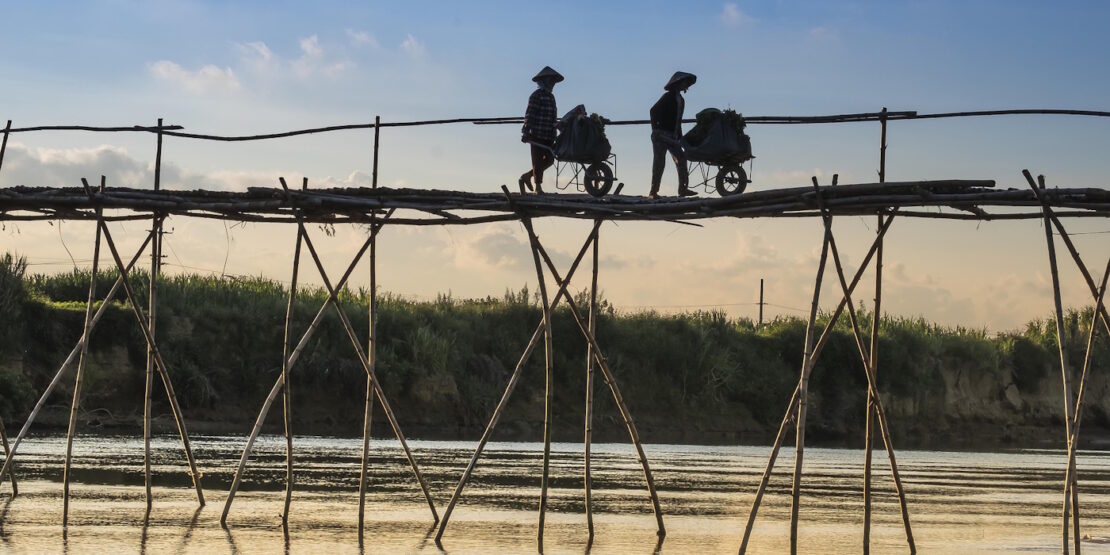 Das Foto zeigt zwei Personen die mit ihren Schubkarren über eine hohe Bambusbrücke gehen