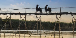 Das Foto zeigt zwei Personen die mit ihren Schubkarren über eine hohe Bambusbrücke gehen