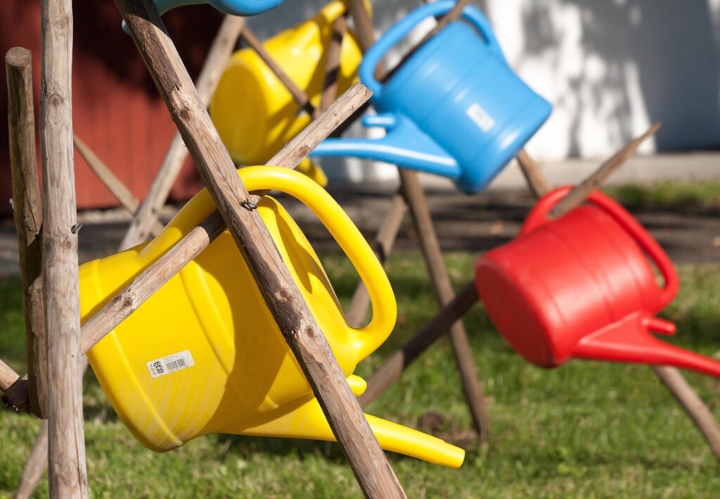 Das Foto zeigt bunte Gießkannen die an Holzpfählen hängen