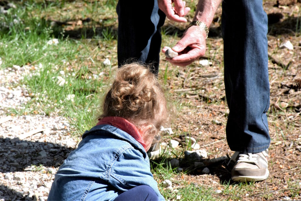 Das Foto zeigt ein Kind, das im Garten sitzt und einen Erwachsenen, der einen Stein hält