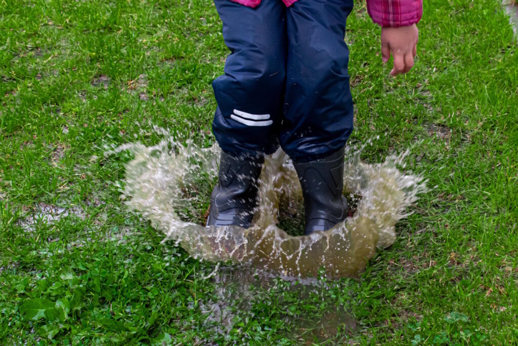 Das Foto zeigt ein Kind mit Matschhose und Gummistiefel, das in eine Pfütze hüpft